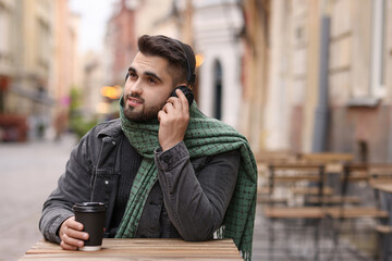 Handsome man in warm scarf with paper cup listening to music in outdoor cafe. Space for text