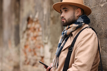 Obraz premium Handsome man in warm scarf with smartphone near wall outdoors. Space for text