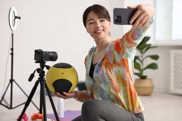 Happy sports blogger holding medicine ball while streaming online fitness lesson with smartphone at home