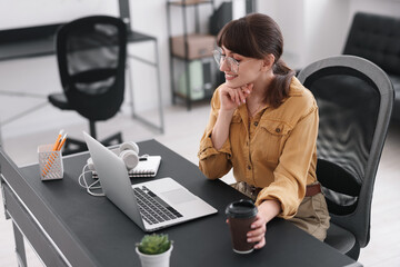 Woman with cup of coffee watching webinar at table in office