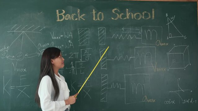 Back to school concept. Asian female teacher smiling with wooden stick pointing to blackboard at school in classroom, Happy beautiful young woman standing hold pointer to back board