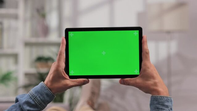 Closeup shot of a man lying on a sofa using a tablet in a horizontal position with green screen mockup in a bright room, tapping on the screen to start a video.