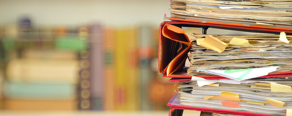stack of messy file folders with narrow depth of field, blurred bookshelf  in the back,red tape,...