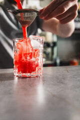 A hand pours a vibrant red beverage through a strainer into a glass with ice cubes.