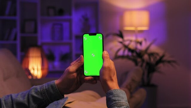Closeup shot of a man lying on a sofa using a smartphone in a vertical position with green screen mockup in a evening room, tapping on the screen to start a video.