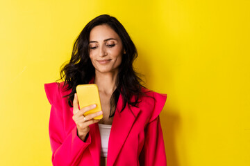 Studio portrait of a surprised young brunette looking at her phone on a bright yellow backgroun