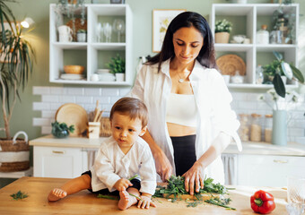 Fototapeta premium A young mother is cutting greens on a wooden stand in a modern kitchen and her little baby is playing next to her on the table.