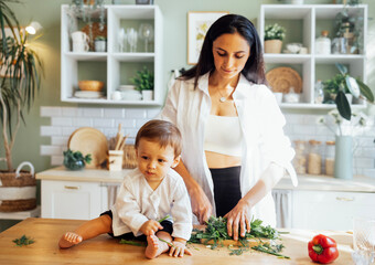 A young mother is cutting greens on a wooden stand in a modern kitchen and her little baby is...