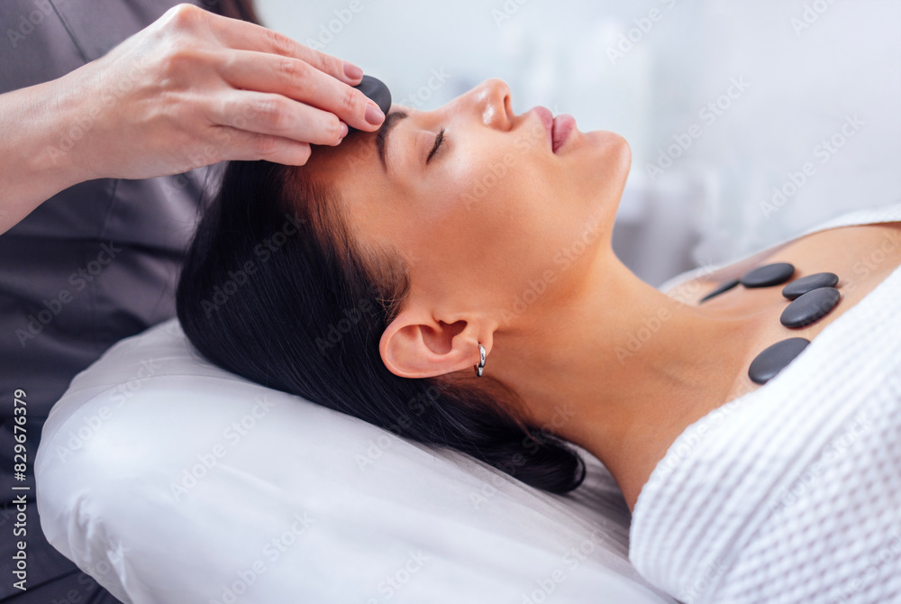 Wall mural close-up of a portrait of a young woman with her eyes closed receiving a facial massage with hot sto