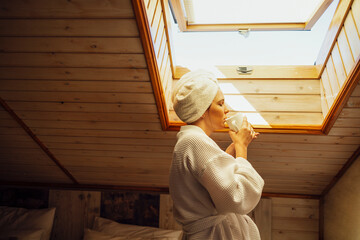 Young woman in a white bathrobe and a towel on her head is holding a cup. Relaxed caucasian female...