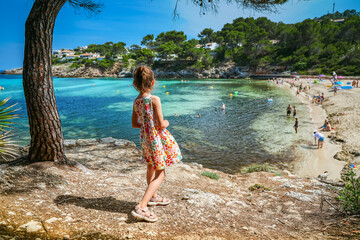 A little girl, standing under a pine tree by the shore, looking out over the clear blue sea and...