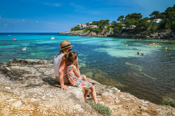 Mother and little daughter sitting under a pine tree by the shore, looking over the clear blue sea....