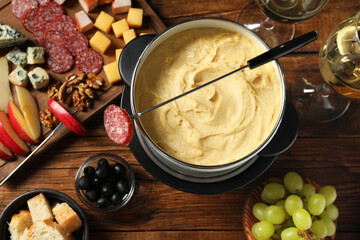 Fork with piece of sausage, melted cheese in fondue pot and other products on wooden table, flat lay