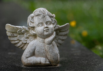 Close-up of a small old and weathered angel standing on a gravestone. Green meadow in the background.
