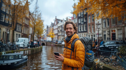 Naklejka premium Smiling man in a yellow jacket holding a phone by a canal with autumn trees and architecture