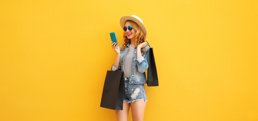Beautiful happy young woman holding phone with black shopping bag, summer hat on yellow background