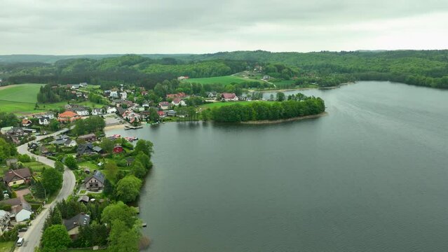 Aerial view of Ostrzyce village by a lake, surrounded by lush green hills and forests.