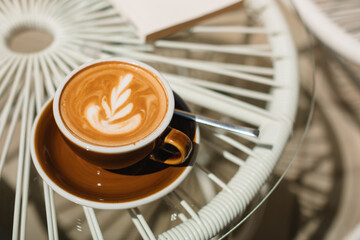 A cup of cappuccino and a book on a glass table on the terrace on a sunny summer morning.