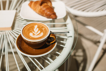 A cup of fragrant cappuccino and a warm croissant on a glass table on the terrace of the cafe.