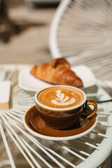 A cup of fragrant cappuccino and a warm croissant on a glass table on the terrace of the cafe.