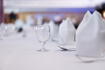 Close-up of a clear wine glass with a traditional round shape. On a white dining table with eating utensils, there is a blurry background of the restaurant