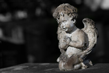 Fototapeta premium Close-up of a small old and weathered angel kneeling on a gravestone. There is space for text. The picture is monochrome
