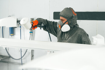 Male painter in respiratory mask and protective suit painting car with spray gun in car service
