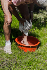 An adult elderly man gardener washes his dirty feet in a red basin, a bowl of water on the green...