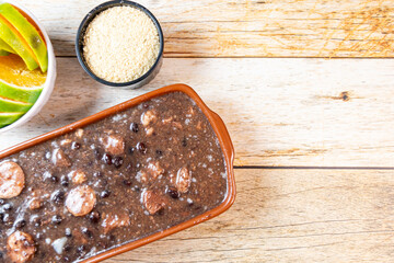 Traditional Brazilian feijoada with black beans, sausage, and farofa served with fresh orange slices on a rustic wooden table.