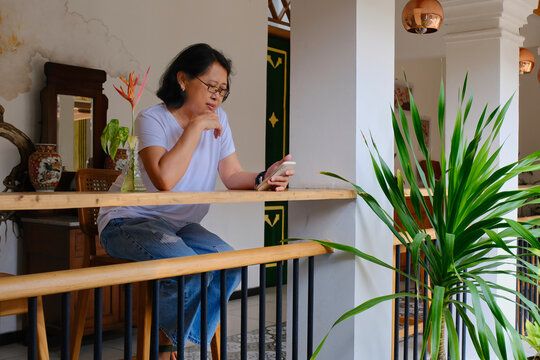 Asian woman sitting in her home veranda reading messages on her smartphone