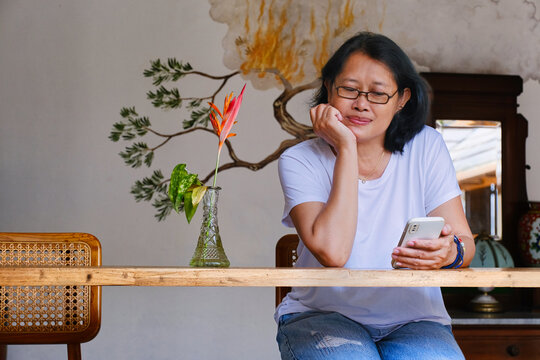 Asian woman sitting alone on her home veranda reading messages on her smartphone