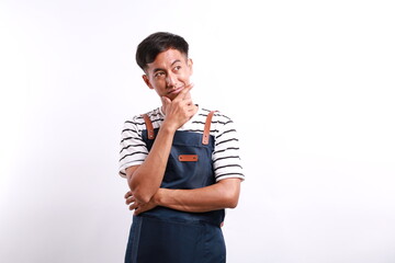 Thoughtful Asian man in apron keeps hand on chin looks pensively above against white background...
