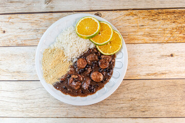 Traditional Brazilian feijoada, a hearty black bean stew with sausages, served with rice, orange slices, and farofa on a rustic wooden table.