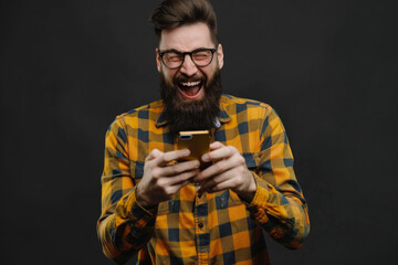 Exuberant man cheering and clenching fists in victory while looking at his smartphone against a neutral background.