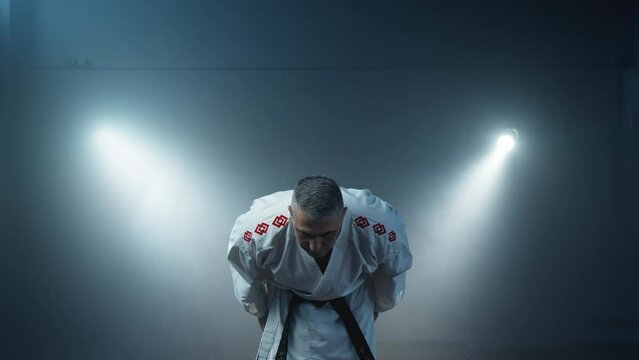 Focused martial artist in white kimono with black belt bows in dimly lit dojo. Man shows discipline and dedication to the art. Karate fighter at foggy background. 4K, UHD