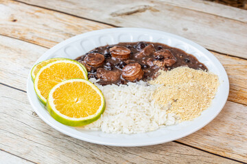 Traditional Brazilian feijoada served with white rice, orange slices, and farofa on a rustic wooden table.
