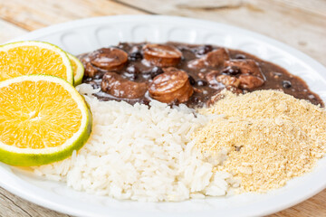 Traditional Brazilian feijoada served with white rice, orange slices, and farofa on a white plate. A delicious and hearty meal.