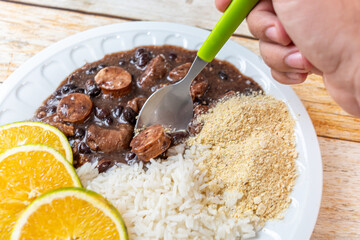 A traditional Brazilian feijoada served with black beans, sausages, rice, cassava flour, and orange slices on a white plate.