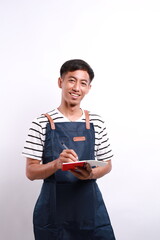 Smiling young Asian man barista barman employee wearing brown apron working in coffee shop write...