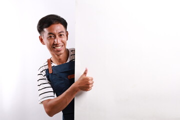 Cheerful Asian man in apron showing thumbs up with something on white advertisement board on white...