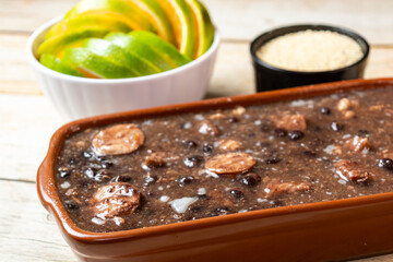 Traditional Brazilian feijoada with black beans and sausage in a clay pot, served with rice and orange slices.