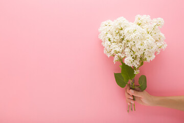 Young adult woman hand holding fresh beautiful bouquet of white lilac flowers with green leaves on light pink wall background. Pastel color. Closeup. Front view. Empty place for text.