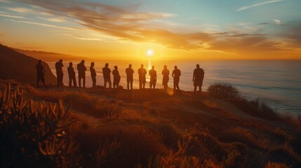 A group of people are standing on a hill overlooking the ocean. The sun is setting, casting a warm glow over the scene. The people are all looking out at the water, taking in the beauty of the moment