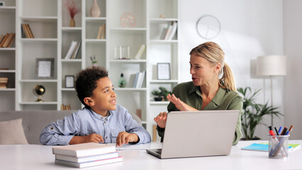 Caring female tutor helping little African American elementary school student with homework at home while sitting at the table. Happy boy doing homework on laptop, studying at home with babysitter.