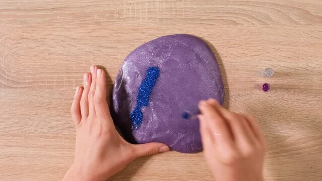 Slime DIY concept. Female hands sprinkle colorful glitter on the slime. 4k resolution video of close up top view of female hand holding purple shining slime and squeezing it.