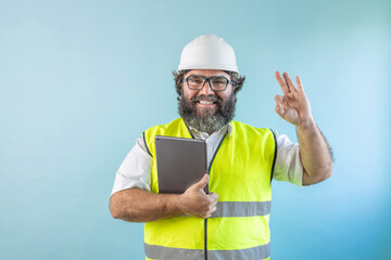 smiling adult man engineer with beard and wearing helmet and safety vest looking at camera on blue background, using his tablet and reviewing work, pointing at something or someone