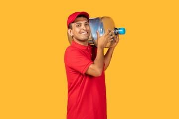 A cheerful young delivery man in a red uniform and cap holds a large water container on his...