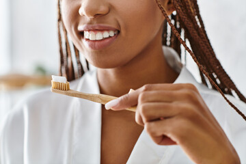 An African American woman in a bathrobe with afro braids brushes her teeth in a modern bathroom for...