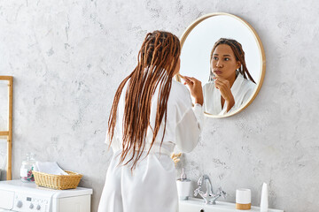 An African American woman with afro braids stands in her modern bathroom, brushing her teeth in...