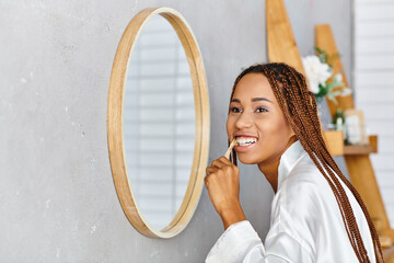 An African American woman with afro braids in a bathrobe brushing her teeth in front of a mirror in...